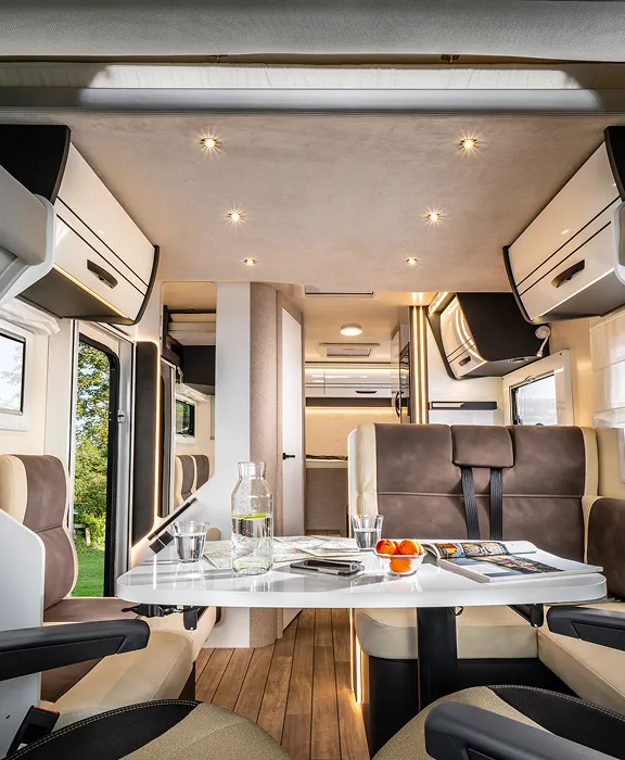 Blick aus dem Fahrerhaus in den modernen, hellen Wohnbereich eines Wohnmobils mit Sitzgruppe, Tisch und Hubbett.