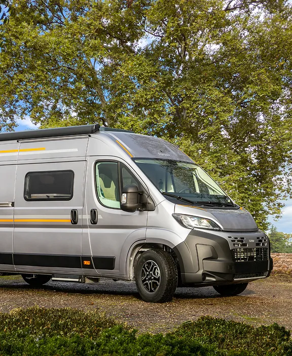 Silberner Forster Van mit Markise und gelben Akzenten parkt auf einem Waldweg vor grünen Bäumen.